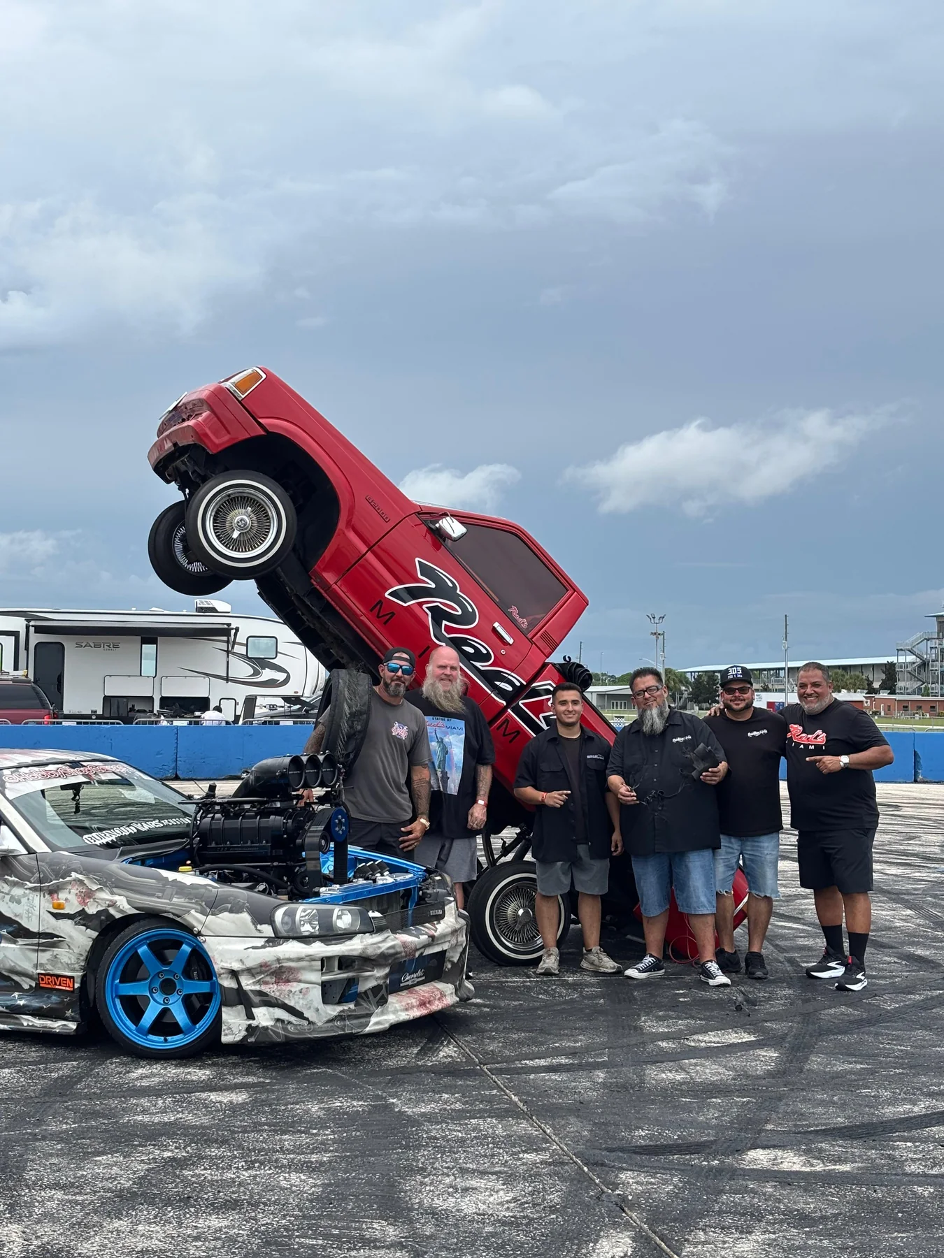 Six men in Red's Miami shirts standing beside a red Red's pickup lifted nearly vertical on hydraulics at an outdoor event, with a wrapped, supercharged car in the foreground