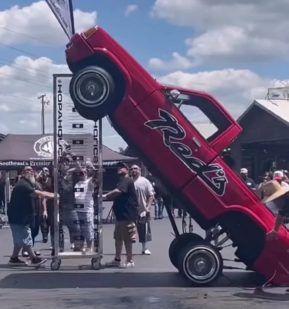Red Red's pickup lifted steeply on hydraulics at an outdoor automotive event, with spectators and vendor tents behind it