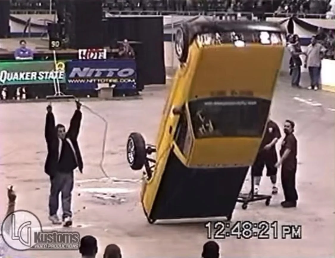 Yellow custom car tipping past vertical on hydraulics at an indoor car show as a spectator raises both arms nearby