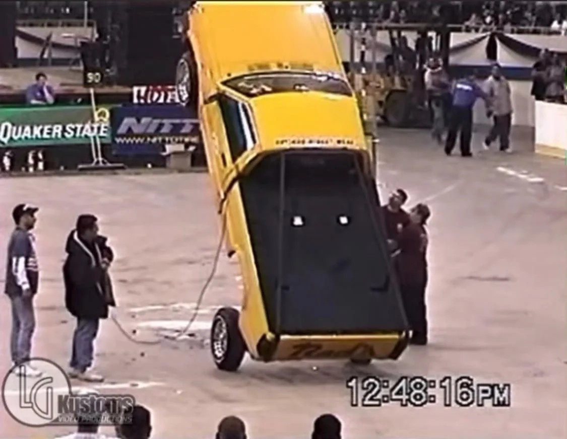 Yellow custom car lifted steeply onto its rear wheels on hydraulics at an indoor car show, with onlookers standing nearby