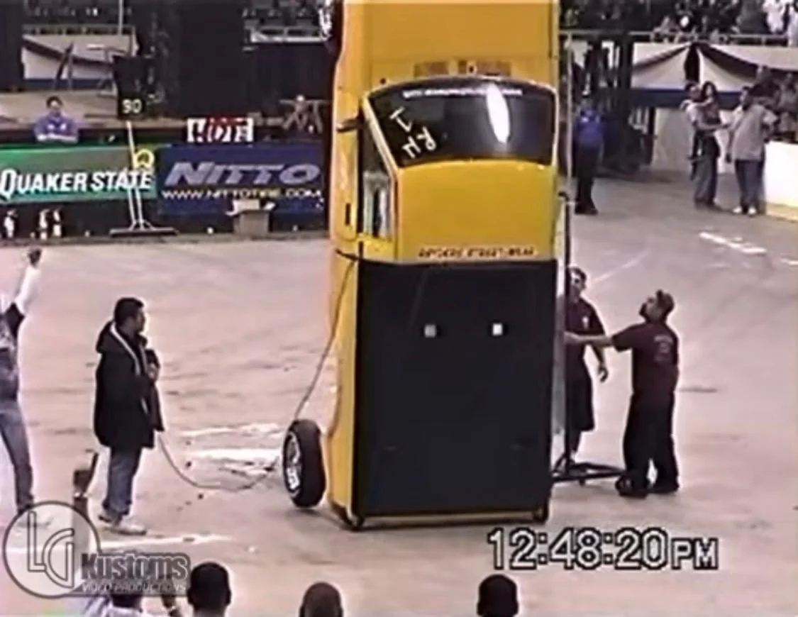 Yellow custom car standing fully vertical on its rear end on hydraulics at an indoor car show while crew members watch from the floor